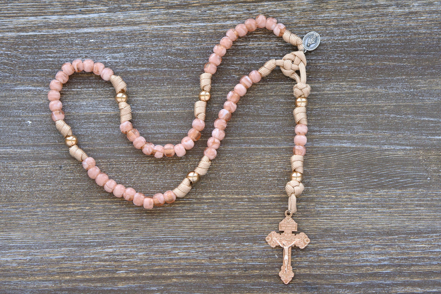 A child's paracord rosary, "The Gate of Heaven," features rose-gold and tan paracord interwoven with milky pink Hail Mary beads and shiny gold Our Father beads. It includes a unique two-inch rose-gold Pardon crucifix and a smaller Miraculous Medal devotional medal.