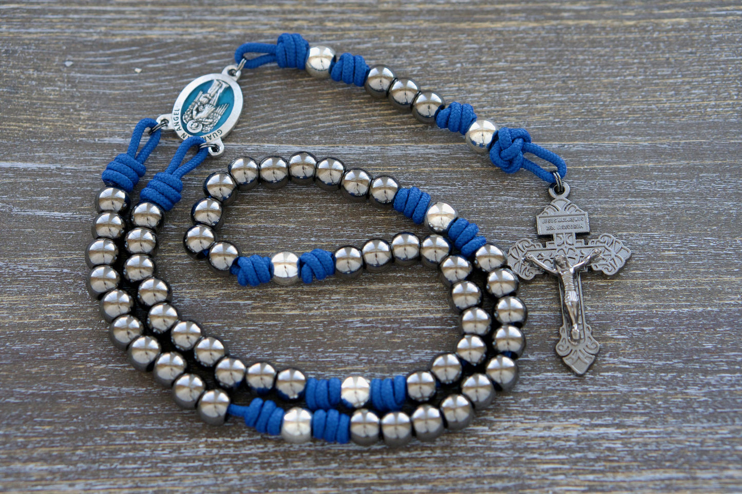 Guardian of Grace Rosary: A premium paracord rosary featuring a striking blue enamel Guardian Angel centerpiece. Durable royal blue paracord, gunmetal and silver metal alloy beads, and a 2-inch gunmetal pardon crucifix make this rosary both elegant and resilient.