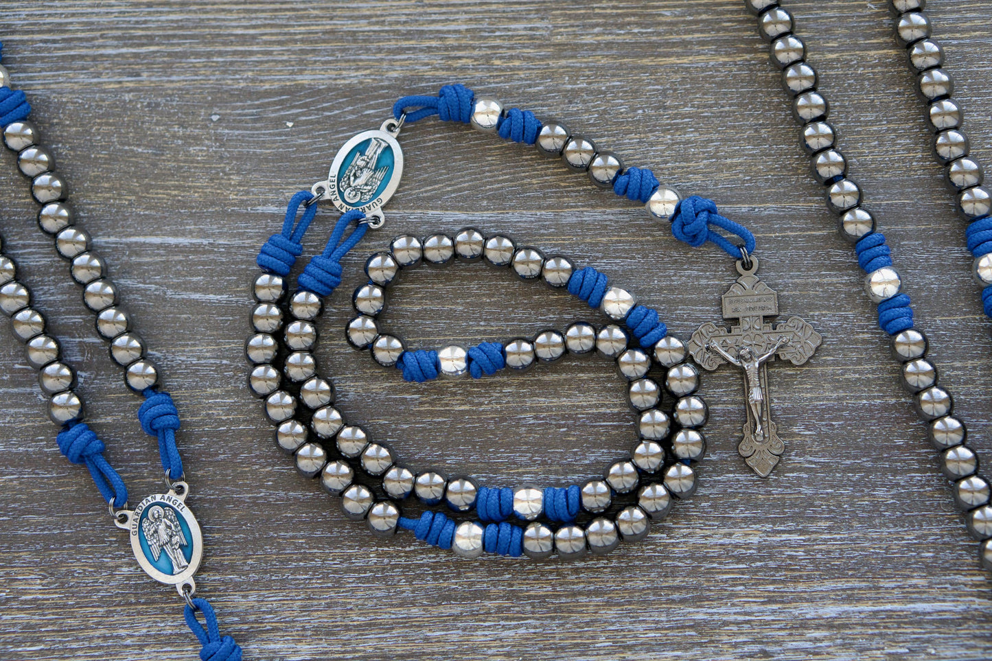 Premium metal paracord rosary featuring a blue enamel Guardian Angel centerpiece, gunmetal Hail Mary beads, silver Our Father beads, and a sturdy 2-inch gunmetal pardon crucifix.  Crafted for durability and devotion, it celebrates the enduring protection and guidance of our Holy Guardian Angels.