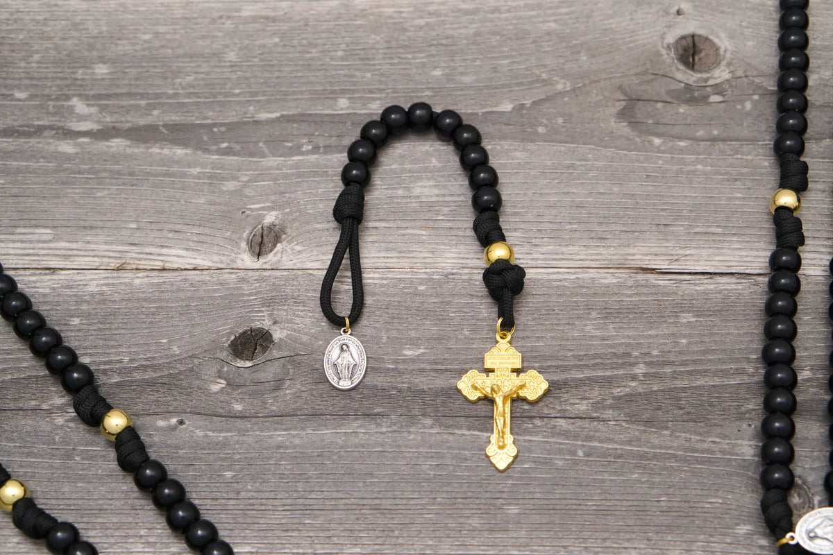 The Holy Servant Paracord Rosary features black beads, gold accents, a Miraculous Medal, and a gold Pardon crucifix.