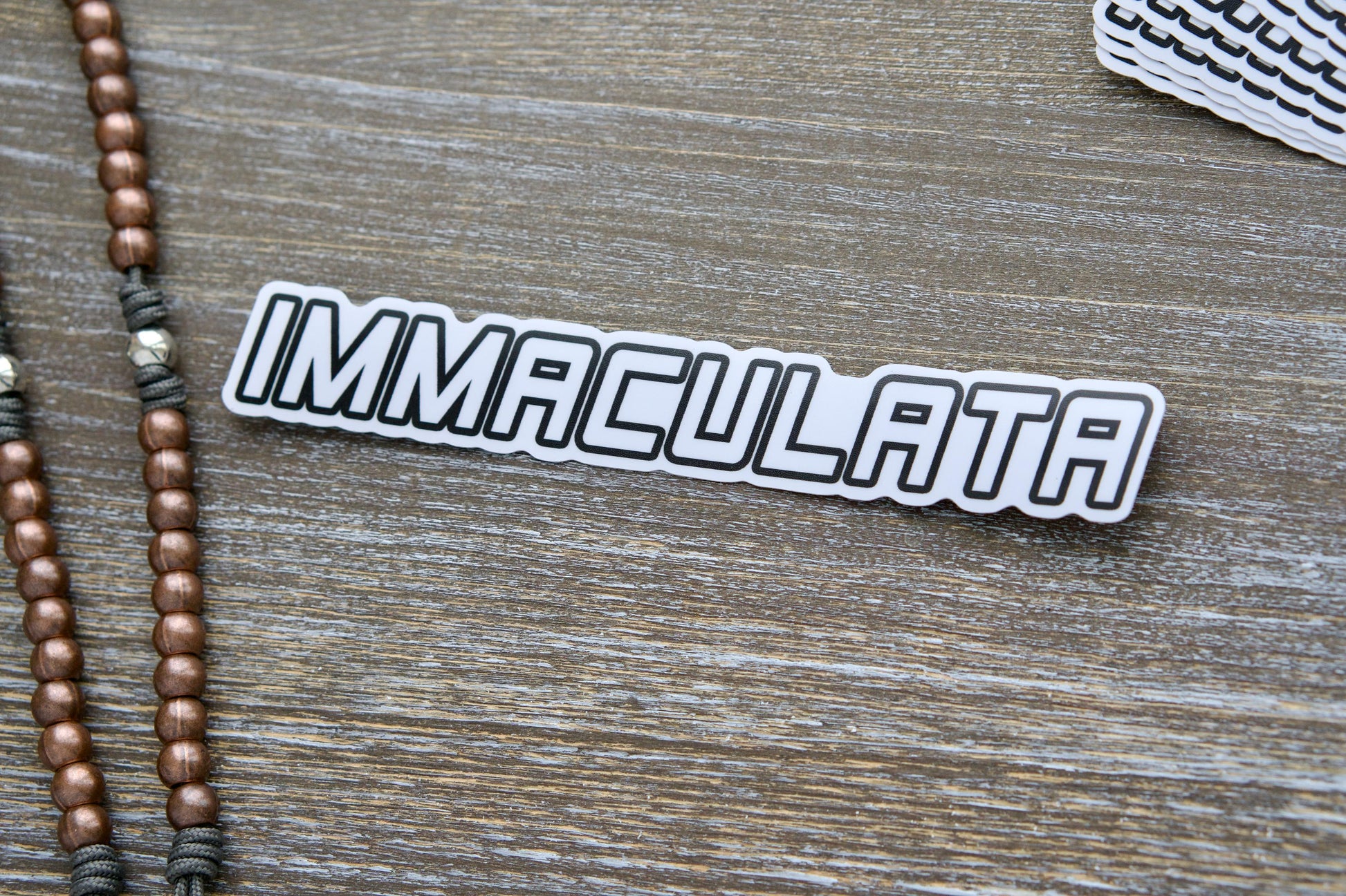 Immaculata - Vinyl Sticker depicting St. Maximilian Kolbe's Militia of the Immaculata emblem for Catholic faith expression.  Removable, UV-resistant, waterproof sticker suitable for indoor/outdoor use, measures 7.17" wide x 1.14" tall.