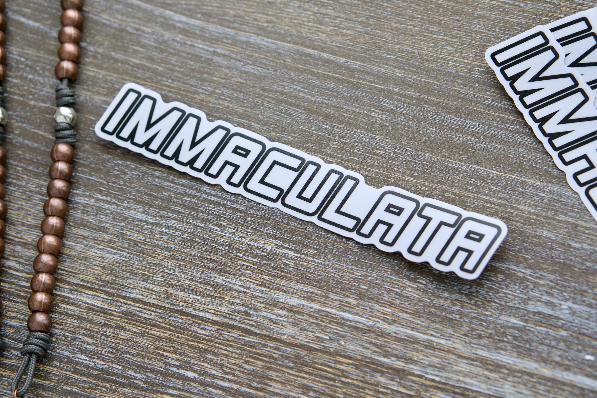 Immaculata Vinyl Sticker - A sleek white vinyl sticker featuring the Militia of the Immaculata emblem. Inspired by St. Maximilian Kolbe's legacy and mission against heresy & modernism. Measures 7.17" wide x 1.14" tall, matte laminated for durability, UV resistant, and suitable for indoor/outdoor use on most surfaces.