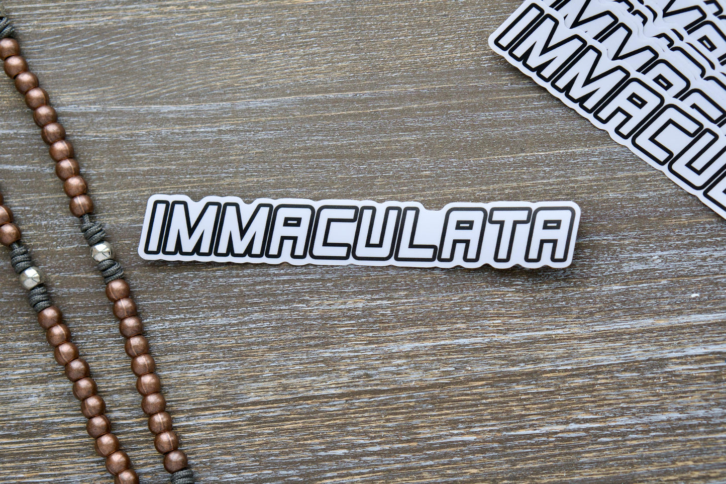 Immaculata Vinyl Sticker featuring the Militia of the Immaculata emblem, inspired by St. Maximilian Kolbe's mission against heresy and modernism. Measures 7.17" wide x 1.14" tall, removable, UV resistant, and suitable for indoor/outdoor use on various surfaces.