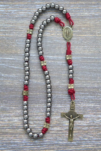 A handcrafted paracord rosary featuring gunmetal Hail Mary beads, an olive Miraculous Medal centerpiece, and a matching St. Benedict crucifix, crimson red rope.