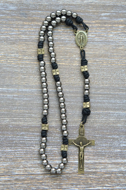 A handcrafted paracord rosary featuring gunmetal Hail Mary beads, an olive Miraculous Medal centerpiece, and a matching St. Benedict crucifix, midnight black rope.