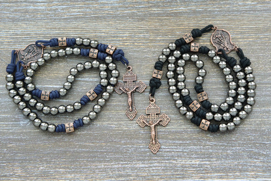 A black or navy blue paracord rosary featuring gunmetal Hail Mary beads, copper St. Michael centerpiece medal, and a crucifix.