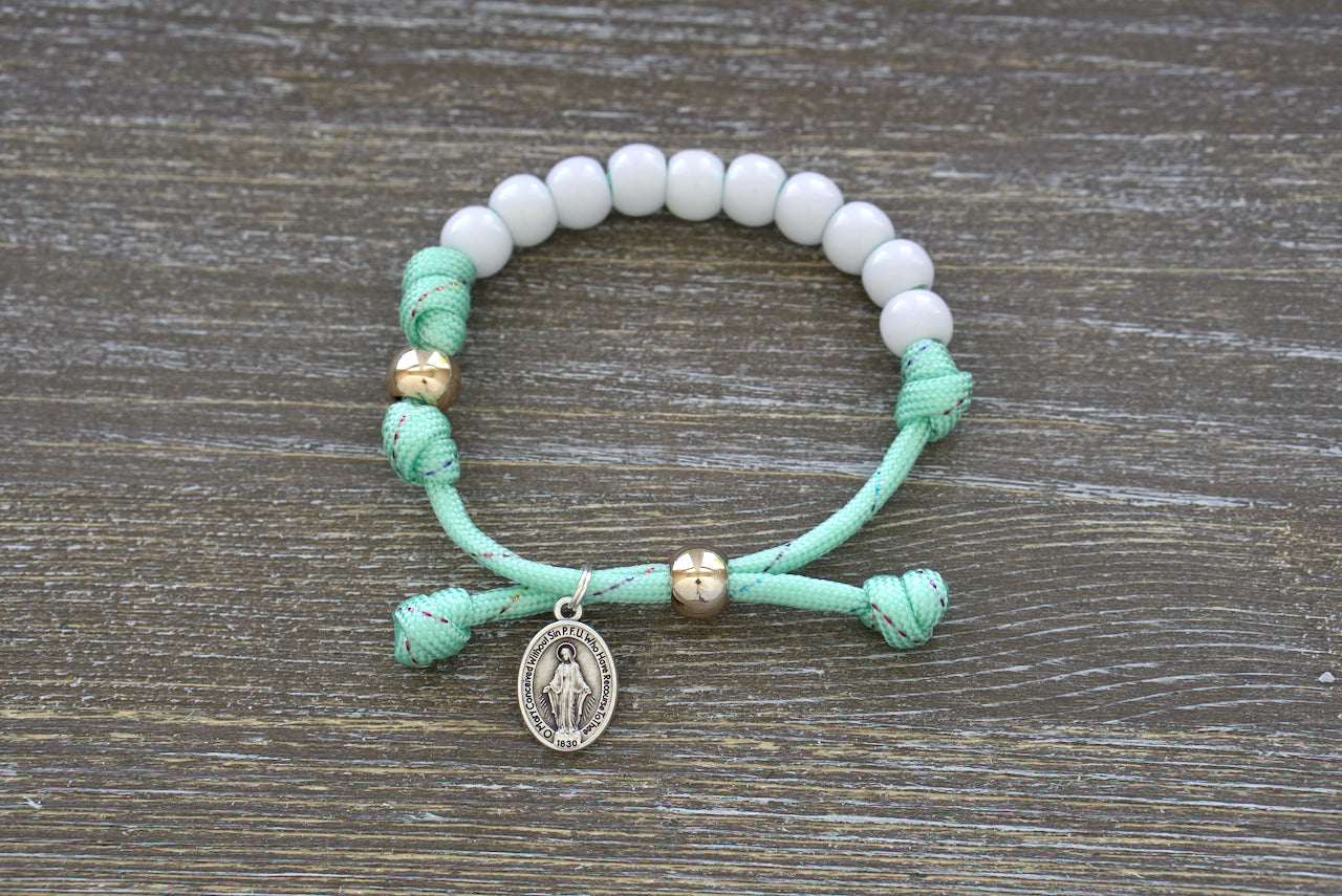 Children's Mint Green Kid's Rosary Bracelet from Sanctus Servo, featuring mint green rope with rainbow ribbon intertwined, white beads for Hail Mary prayers, and a rose gold Our Father bead.