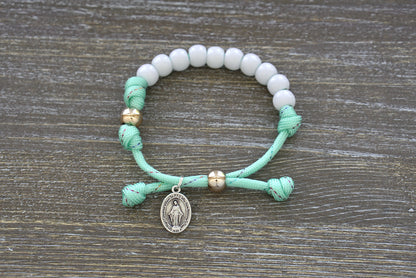 Children's Mint Green Kid's Rosary Bracelet from Sanctus Servo, featuring mint green rope with rainbow ribbon intertwined, white beads for Hail Mary prayers, and a rose gold Our Father bead.