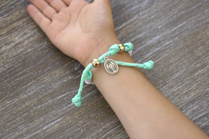 Sanctus Servo Children's Mint Green Kid's Rosary Bracelet: A colorful and handmade rosary bracelet for young Catholics, featuring mint green paracord with rainbow ribbon intertwined, white acrylic beads, a rose gold Our Father bead.