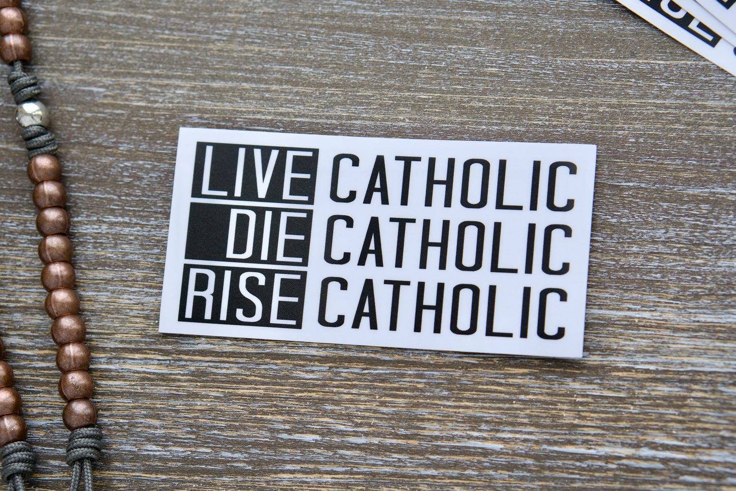 Live Catholic Die Catholic Rise Catholic vinyl sticker design. Featuring a bold text statement inspired by scripture (2 Timothy 4:7-8, John 11:25-26, Revelation 14:13) symbolizing enduring faith, victory over death, and ultimate resurrection. The 4.5" x 2.25" sticker is made from waterproof, durable white vinyl with a semi-glossy finish and matte lamination.