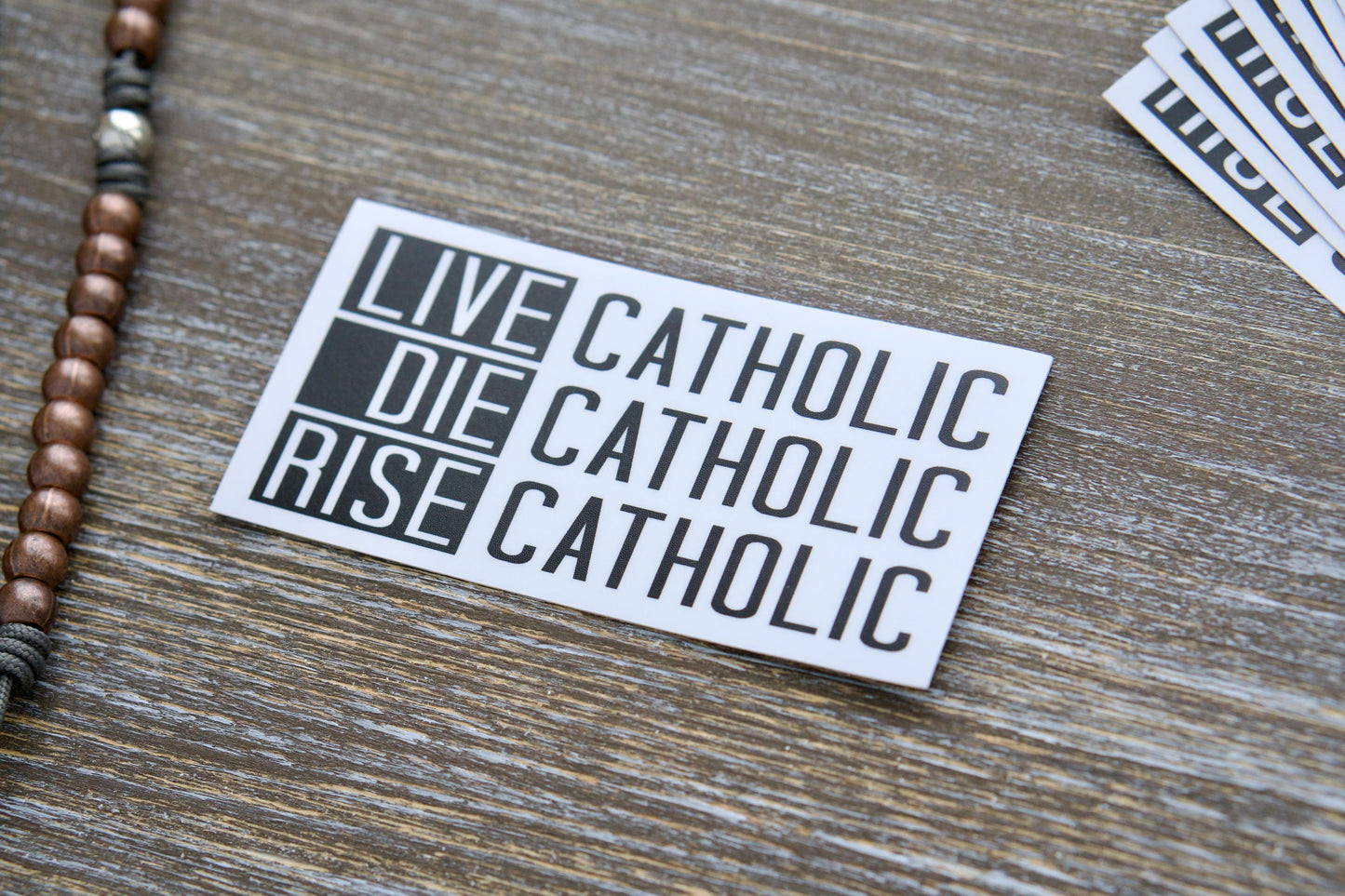 Live Catholic Die Catholic Rise Catholic - Vinyl Sticker featuring bold typography inspired by 2 Timothy 4:7-8, John 11:25-26, and Revelation 14:13. The removable, weatherproof sticker measures 4.5 inches wide by 2.25 inches tall.