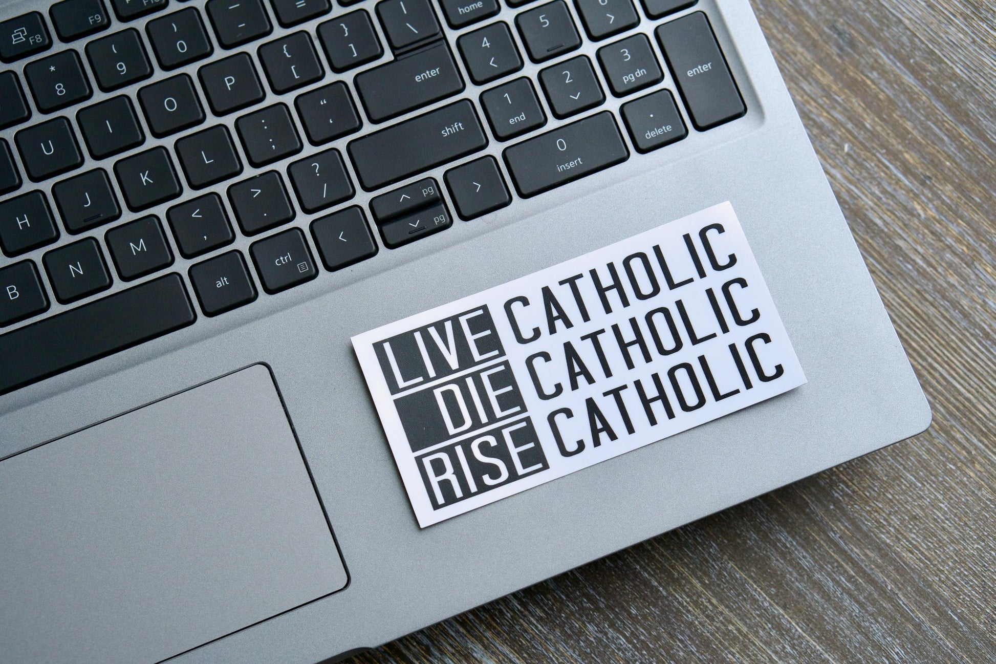 A vibrant white vinyl sticker featuring the bold statement Live Catholic. Die Catholic. Rise Catholic. in an eye-catching font, inspired by 2 Timothy 4:7-8, John 11:25-26, and Revelation 14:13. The sticker measures 4.5" wide x 2.25" tall.