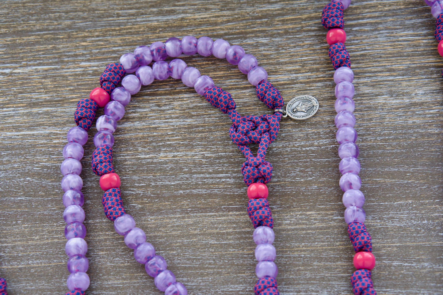 A child's paracord rosary called "Maiden of Faith" featuring milky purple and radiant pink 10mm acrylic beads, accented with a smaller Miraculous Medal and a 2-inch St. Benedict crucifix. The vibrant colors symbolize youthful purity and faith.