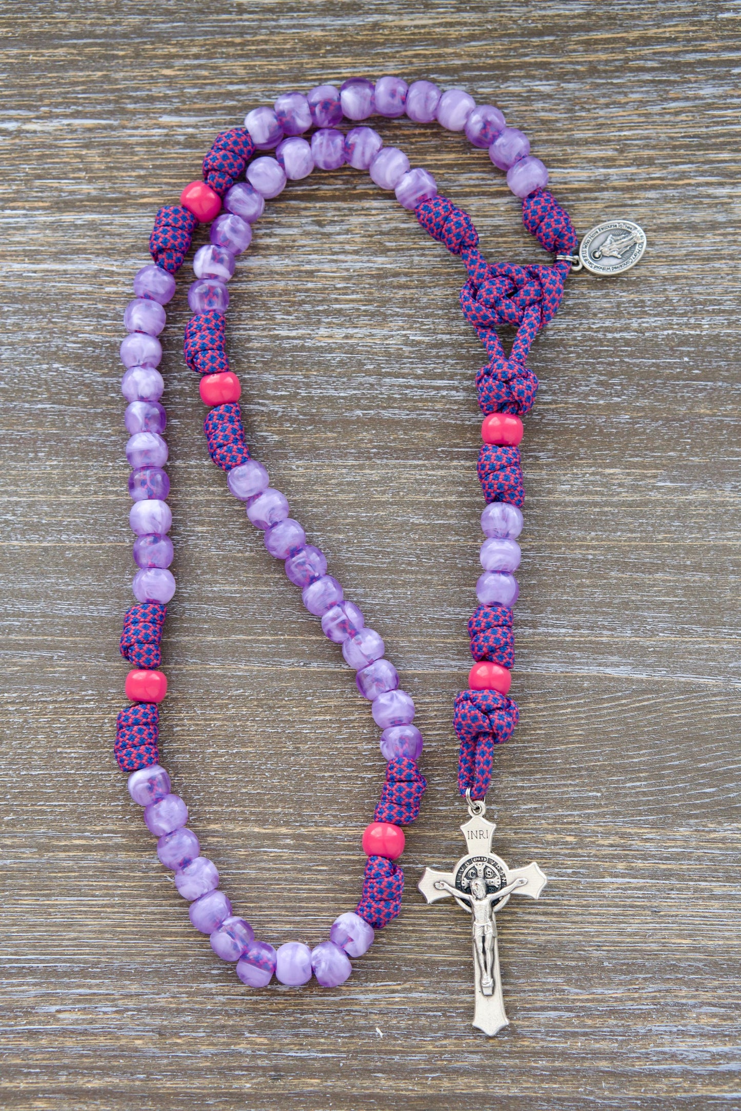 Vibrant pink and milky purple paracord rosary designed for young girls. Features 10mm acrylic beads, a miniature Miraculous Medal, and a 2-inch St. Benedict crucifix. The 'Maiden of Faith' rosary is a durable and symbolic spiritual weapon for Catholic children.