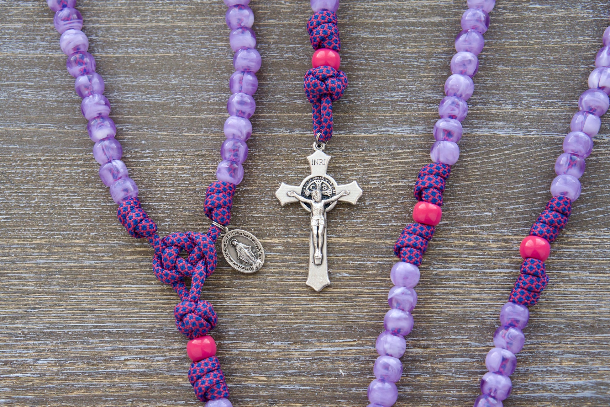 Vibrantly colored paracord rosary designed for children, featuring milky purple and pink 10mm acrylic beads, a smaller 3/4 inch Miraculous Medal, and a 2-inch St. Benedict crucifix. The "Maiden of Faith" rosary is crafted with durable paracord 550.