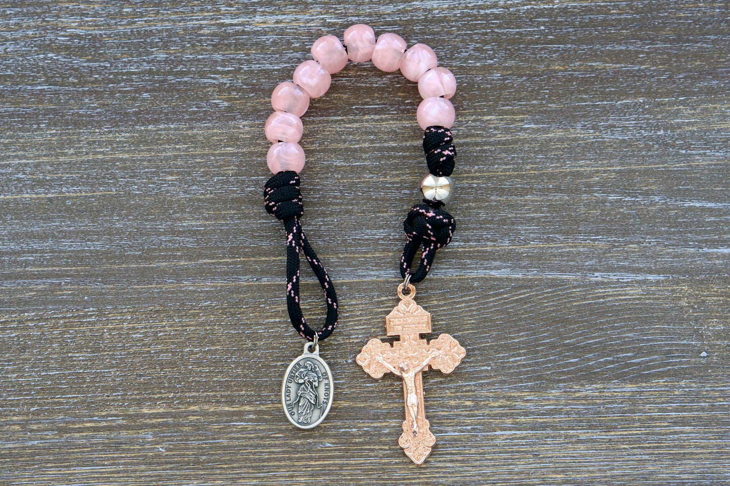 A compact, single decade Our Lady Untier of Knots pocket paracord rosary with pink acrylic Hail Mary beads, a silver Our Father bead, black paracord accented with pink, an 'Our Lady Untier of Knots' medal, and a unique rose gold Pardon crucifix. Handmade by Sanctus Servo.