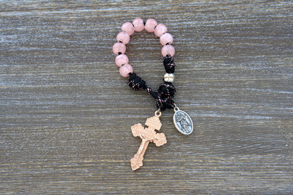 A close-up image showcasing the Our Lady Untier of Knots pocket paracord rosary. Featuring pink acrylic Hail Mary beads, a silver Our Father bead, black paracord with pink accents, and unique devotional medal and rose gold pardon crucifix.