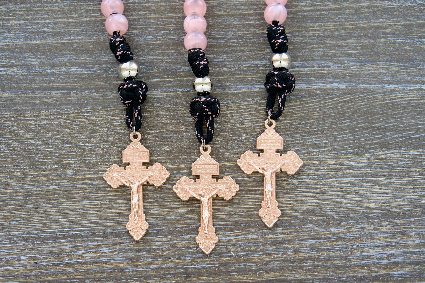 A close-up image of a single decade Our Lady Untier of Knots pocket paracord rosary. It features pink acrylic Hail Mary beads, a shiny silver Our Father bead, black paracord with a pink accent stripe, an Our Lady Untier of Knots devotional medal, and a unique rose gold pardon crucifix.