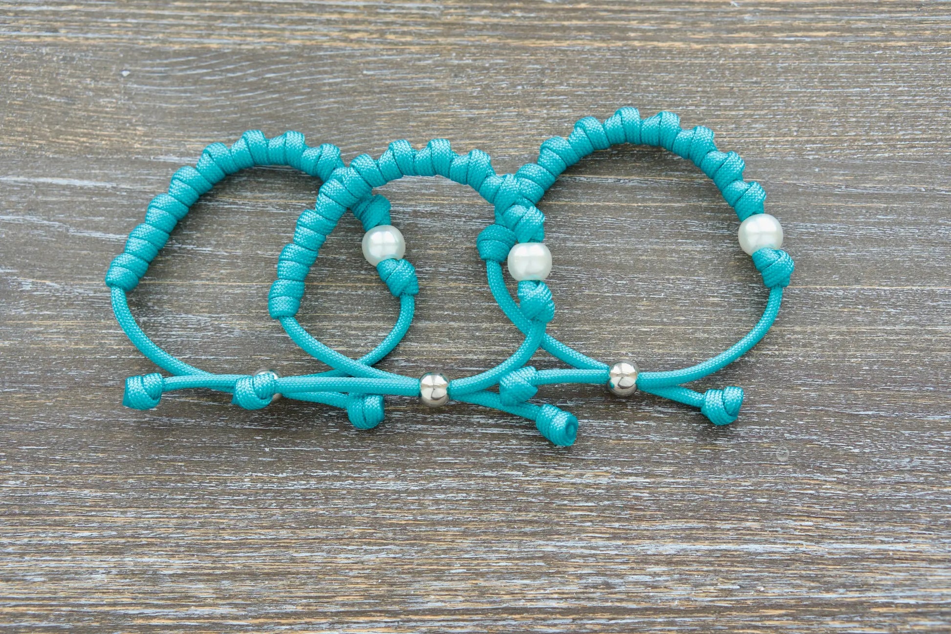 The Peaceful Waters rosary bracelet features teal paracord, a white acrylic Our Father bead, a silver tie bead.