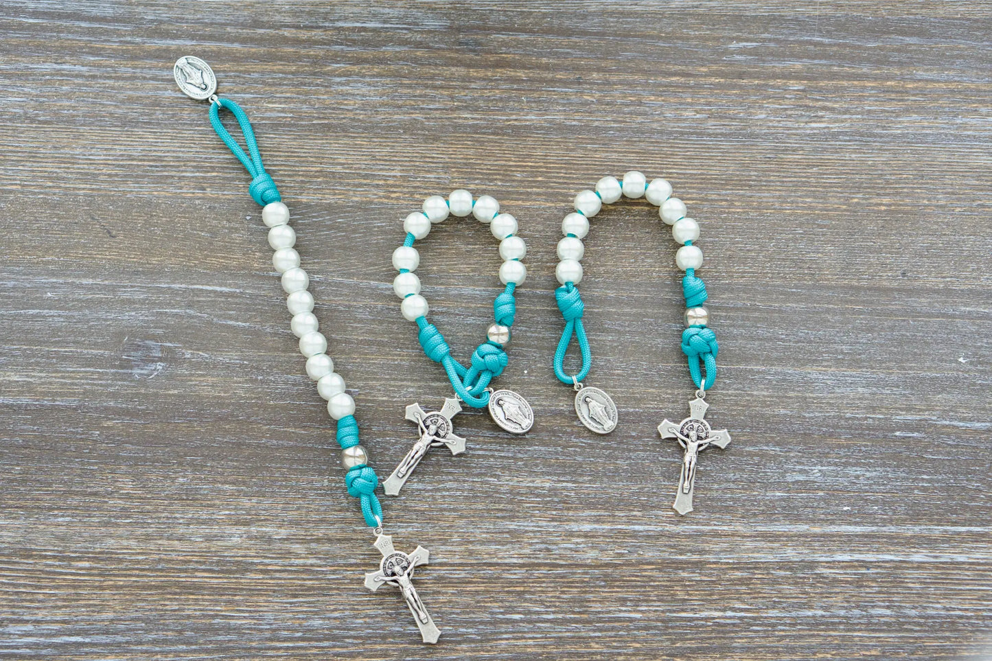 The Peaceful Waters pocket paracord rosary features teal blue 550 cord, pearl white and silver beads, a St. Benedict crucifix, and a Miraculous Medal.
