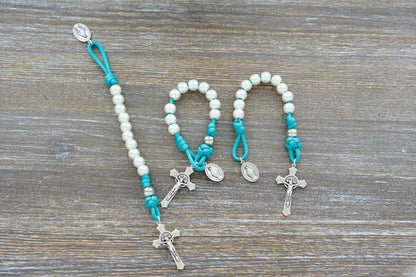 The Peaceful Waters pocket paracord rosary features teal blue 550 cord, pearl white and silver beads, a St. Benedict crucifix, and a Miraculous Medal.