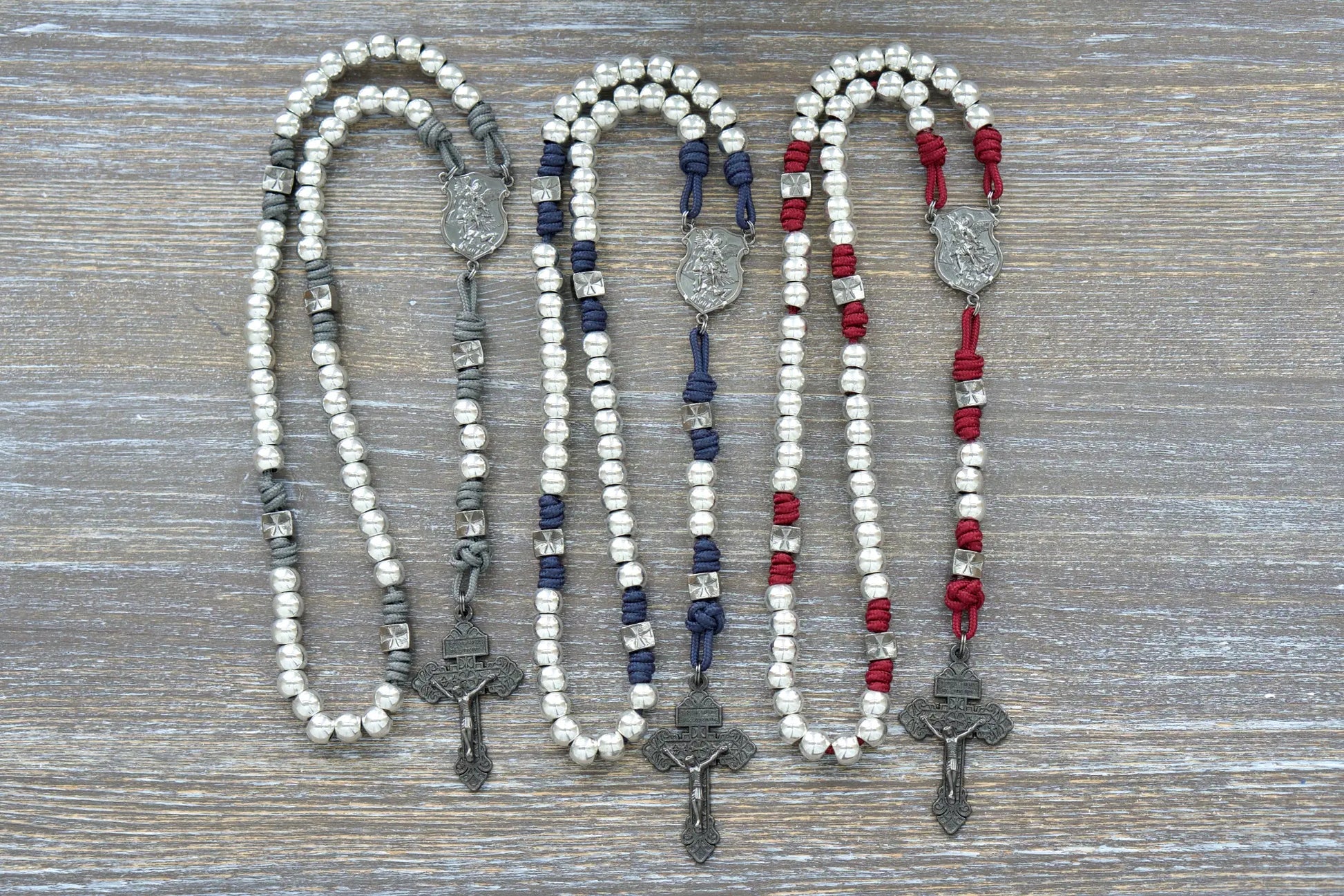 Three rosaries with white, blue, and red paracord rope and silver Hail Mary beads, gunmetal accents.