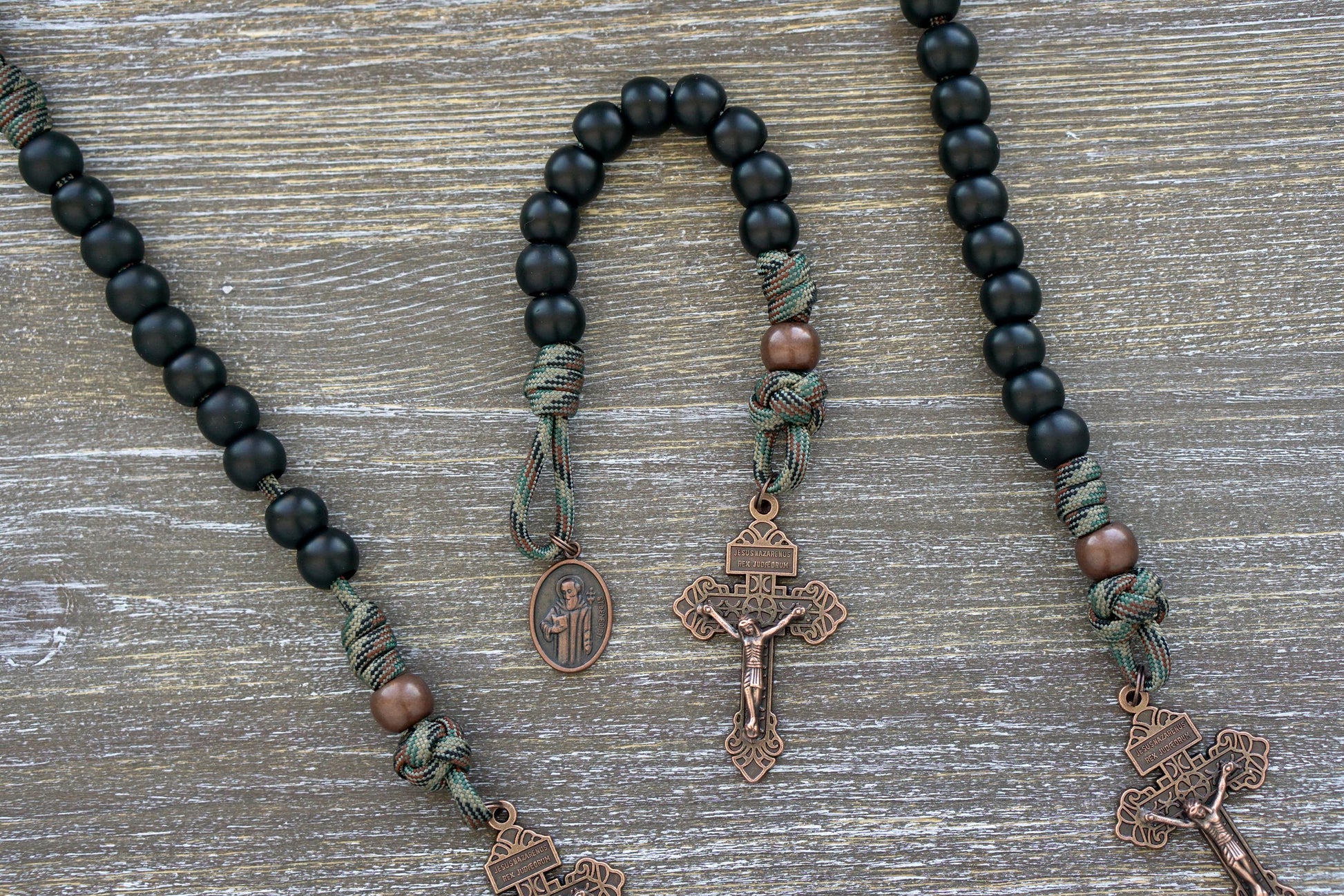 Pocket paracord rosary featuring matte black Hail Mary beads, an antique copper Our Father bead, a St. Benedict medal, and a pardon crucifix. Designed for everyday carry as a symbol of faith and protection against evil.