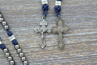Silver crusader Our Father beads and navy blue paracord adorn The Solemn Crusader metal rosary, featuring gunmetal Hail Mary beads, pardon crucifix, and a blue enamel Miraculous Medal.