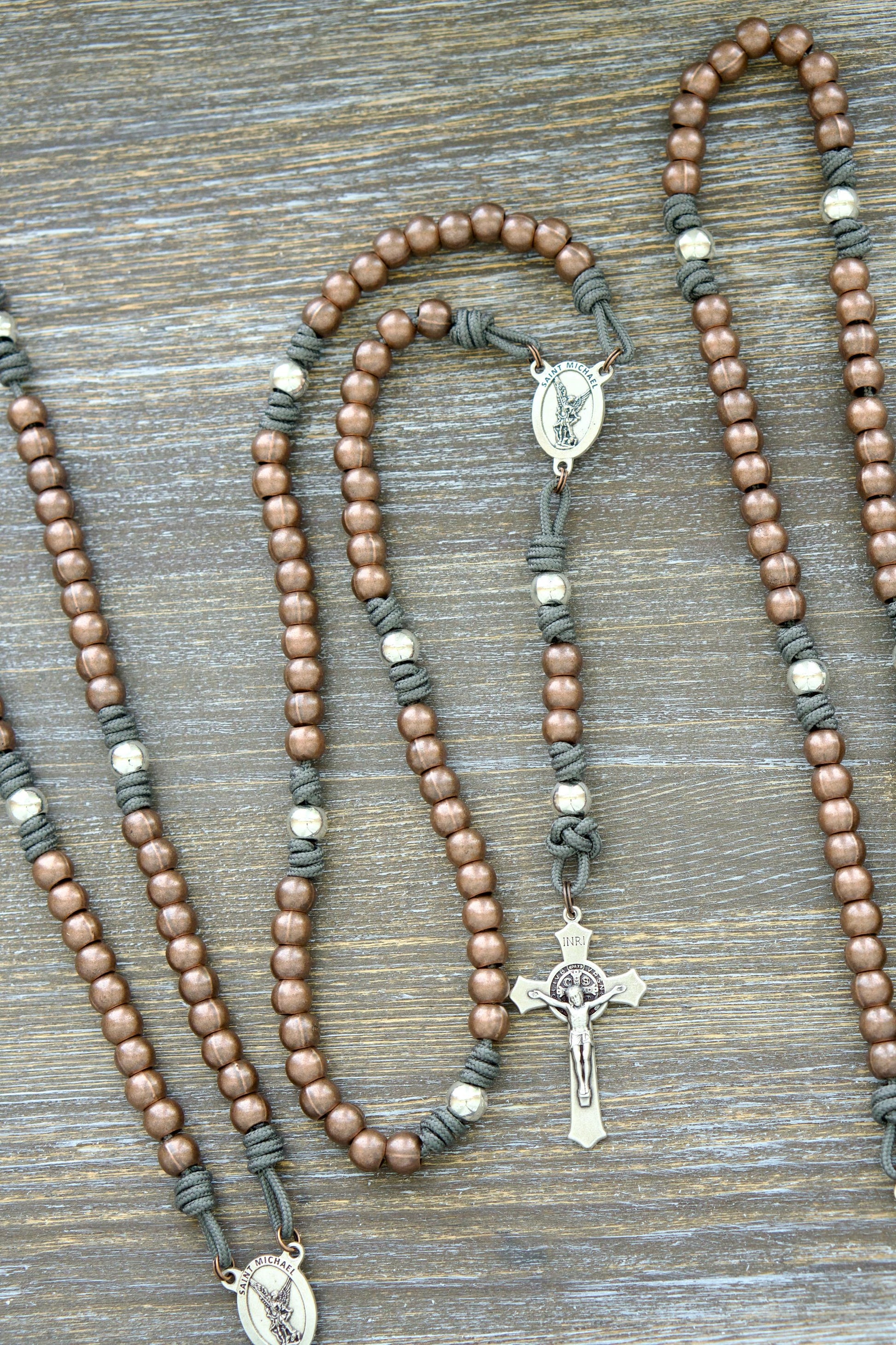 A handcrafted rosary featuring durable paracord (grey, black, green, or navy), silver Our Father beads, copper Hail Mary beads, a radiant St. Michael centerpiece medal, and a 2-inch St. Benedict crucifix.