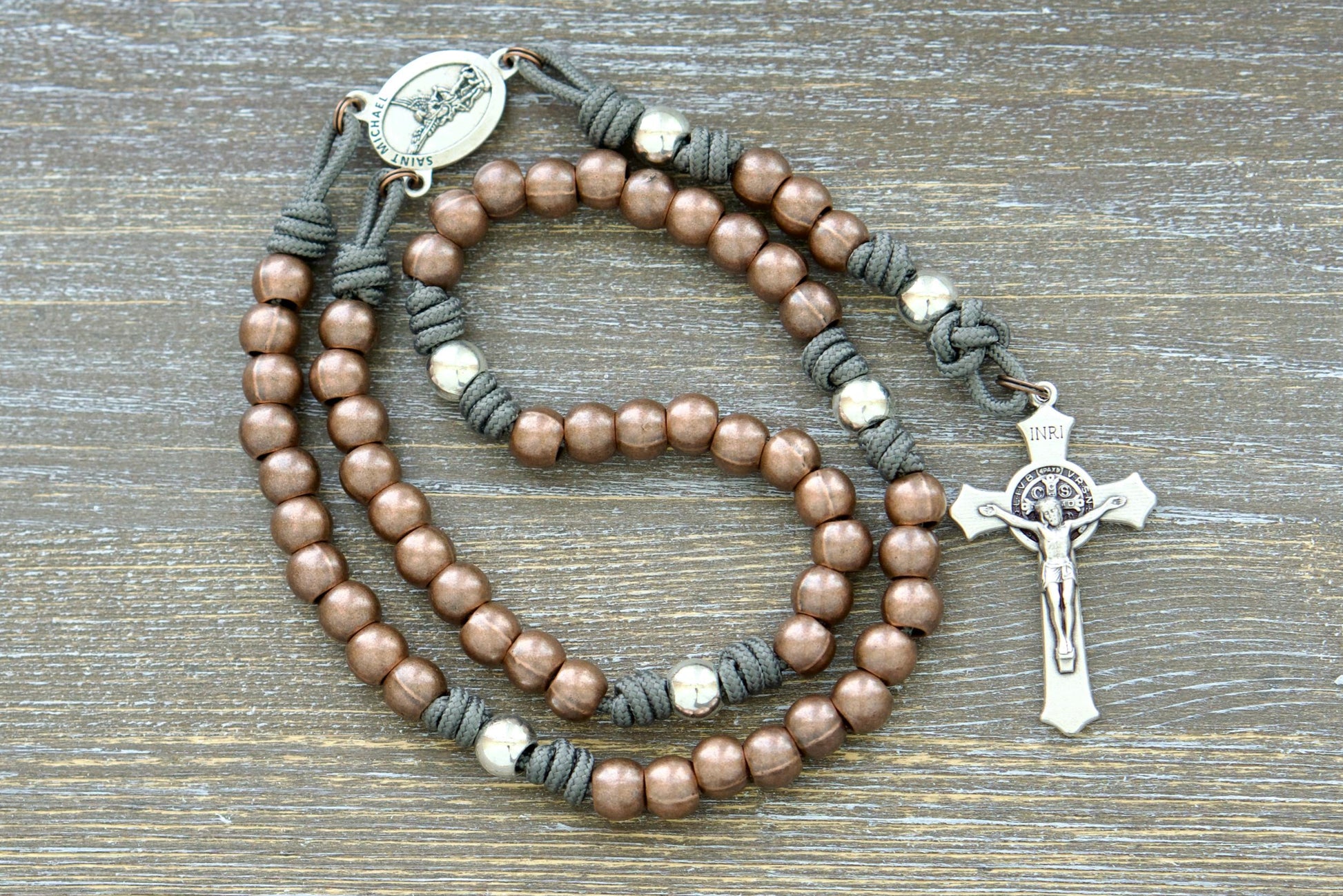  A handcrafted premium paracord rosary featuring durable grey, black, green, or navy blue paracord, 10mm alloy metal Hail Mary (copper) and Our Father (silver) beads, a radiant St. Michael centerpiece medal, and a 2-inch St. Benedict crucifix.