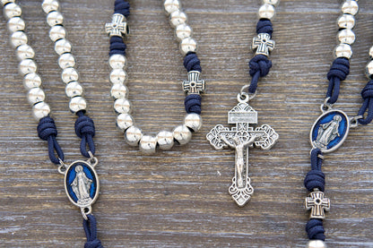 A premium navy blue paracord rosary with shimmering silver metal Hail Mary beads, an intricate cross-shaped Our Father bead design, a blue enamel Miraculous Medal centerpiece, and a matching silver Pardon crucifix. Handmade with durable Paracord.