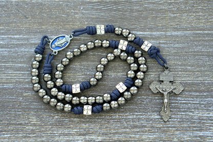 Sturdy navy blue paracord rosary, "The Solemn Crusader," features silver crusader Our Father beads, gunmetal Hail Mary beads, a Miraculous Medal centerpiece, and a 2-inch pardon crucifix.