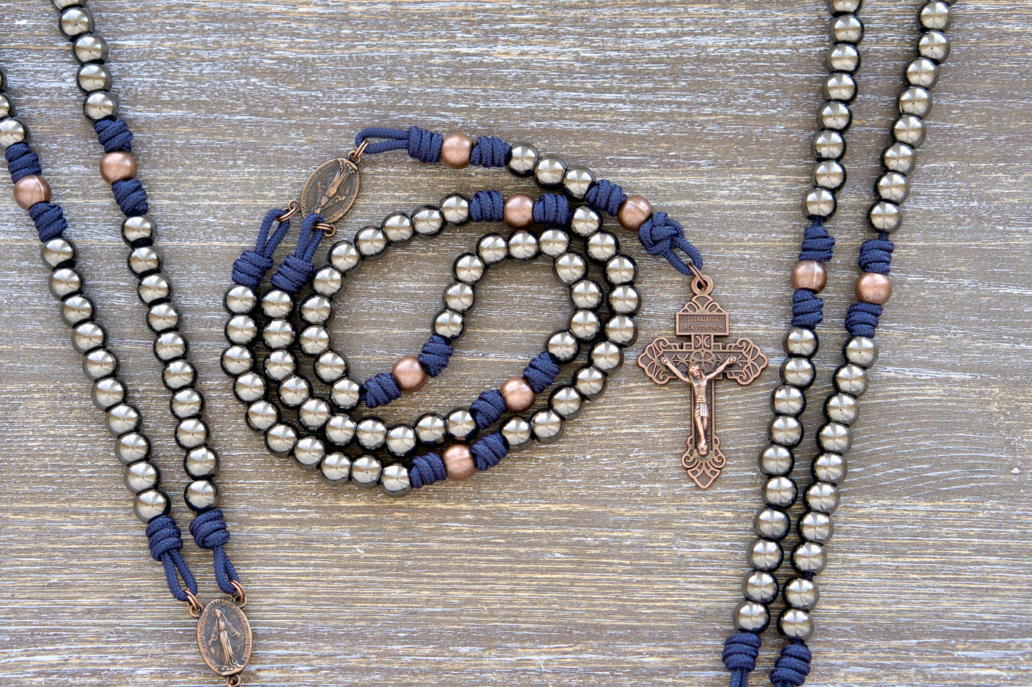 Navy blue paracord rosary featuring antique copper Our Father beads, 10mm gunmetal Hail Mary beads, a copper Miraculous Medal centerpiece, and a copper Pardon crucifix. Designed for durability and everyday carry.