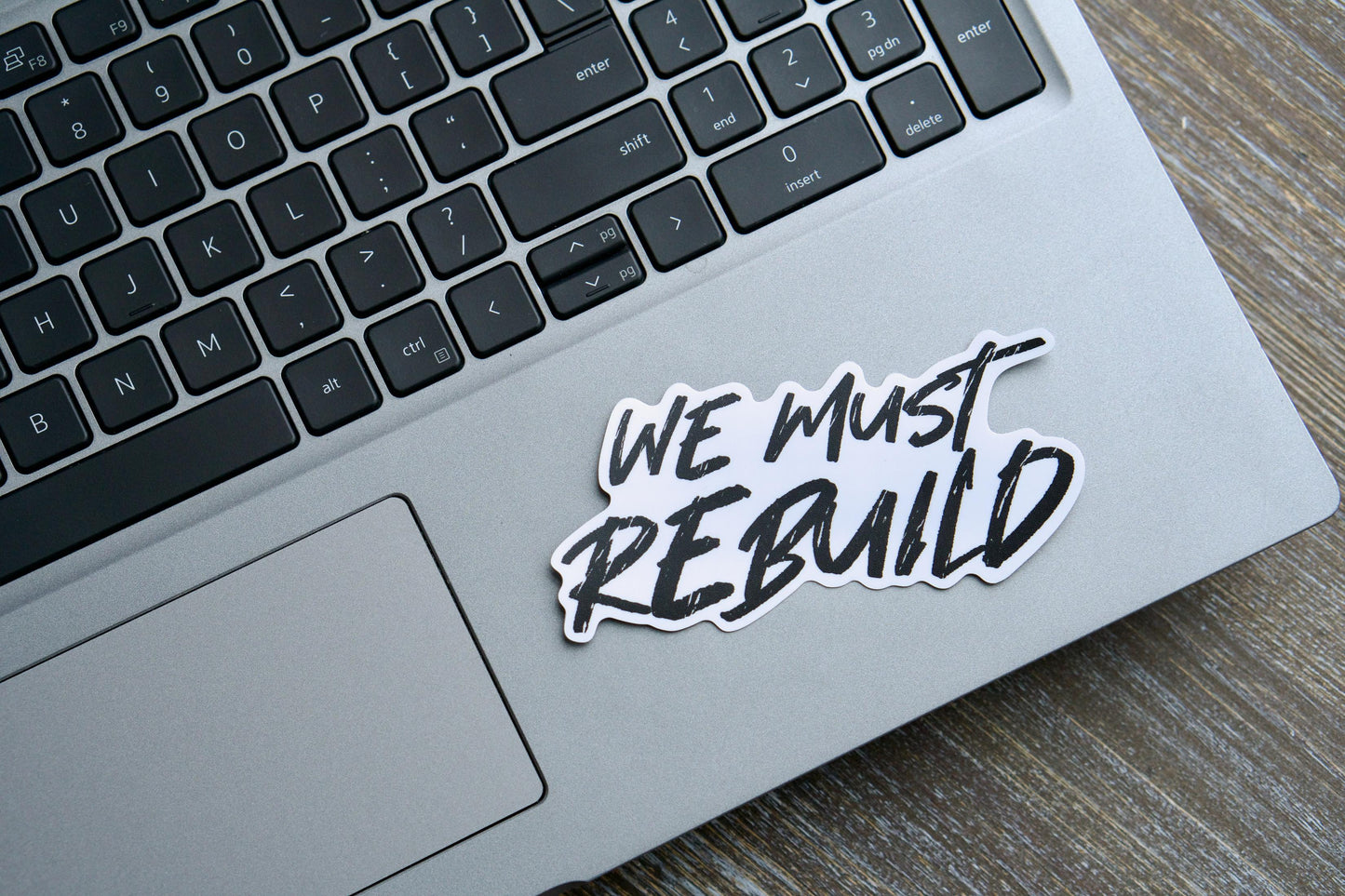 We Must Rebuild - Vinyl Sticker: A 4.25" x 2.13" removable white vinyl sticker with a semi-glossy finish and matte laminate for durability. Features the powerful message "We must rebuild" inspired by Isaiah 61:4