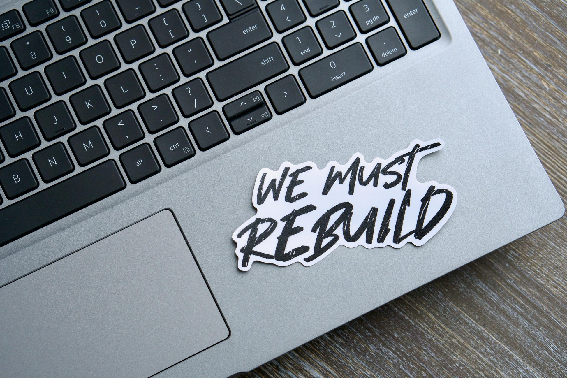 We Must Rebuild - Vinyl Sticker: A 4.25" x 2.13" removable white vinyl sticker with a semi-glossy finish and matte laminate for durability. Features the powerful message "We must rebuild" inspired by Isaiah 61:4