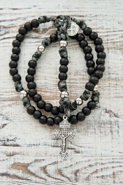 A 5 decade paracord rosary with matte black Hail Mary beads, silver Our Father beads, silver Pardon Crucifix, and black and grey rope paired with a St. Benedict devotional medal.
