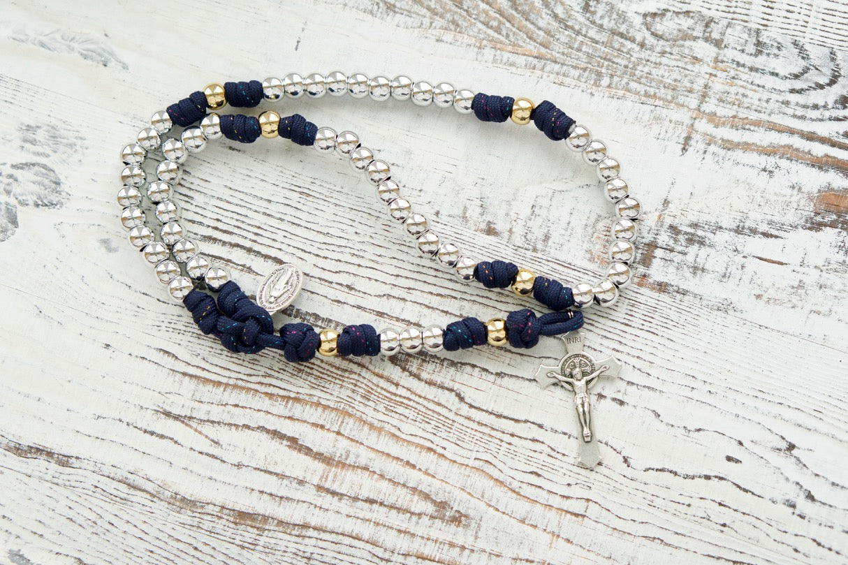 Dark blue paracord rosary with silver Hail Mary beads and gold Our Father beads, featuring a 2" St. Benedict Crucifix and Miraculous Medal. Handcrafted by a small Catholic family for spiritual strength and devotion.