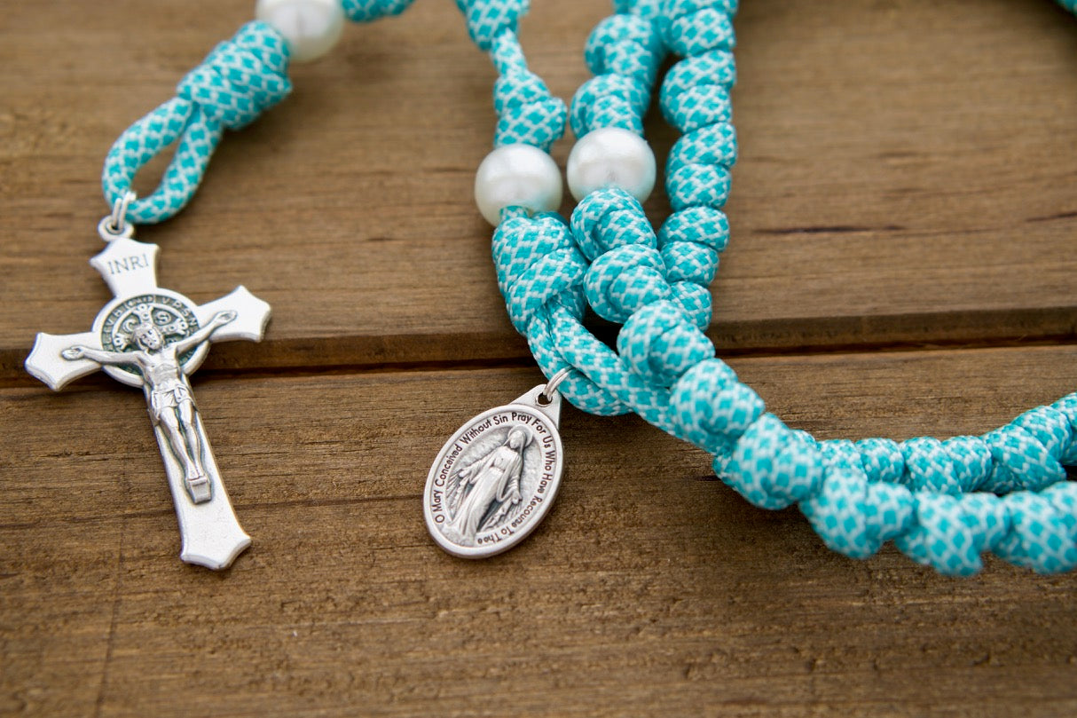 The Peaceful Waters - Teal Blue and White Knotted Rope Rosary, a durable and unbreakable 19-inch Paracord 550 rosary with white pearl Our Father beads, St. Benedict Crucifix, and Miraculous Medal, perfect for spiritual battles with Mary Queen of Peace. Handcrafted by a small Catholic family of 6, customizable and blessed by a priest for maximum indulgences.